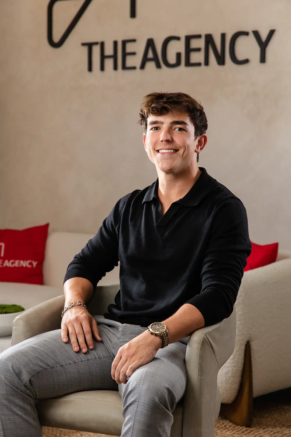 Agent in a black polo seated in a lounge chair beneath a The Agency sign