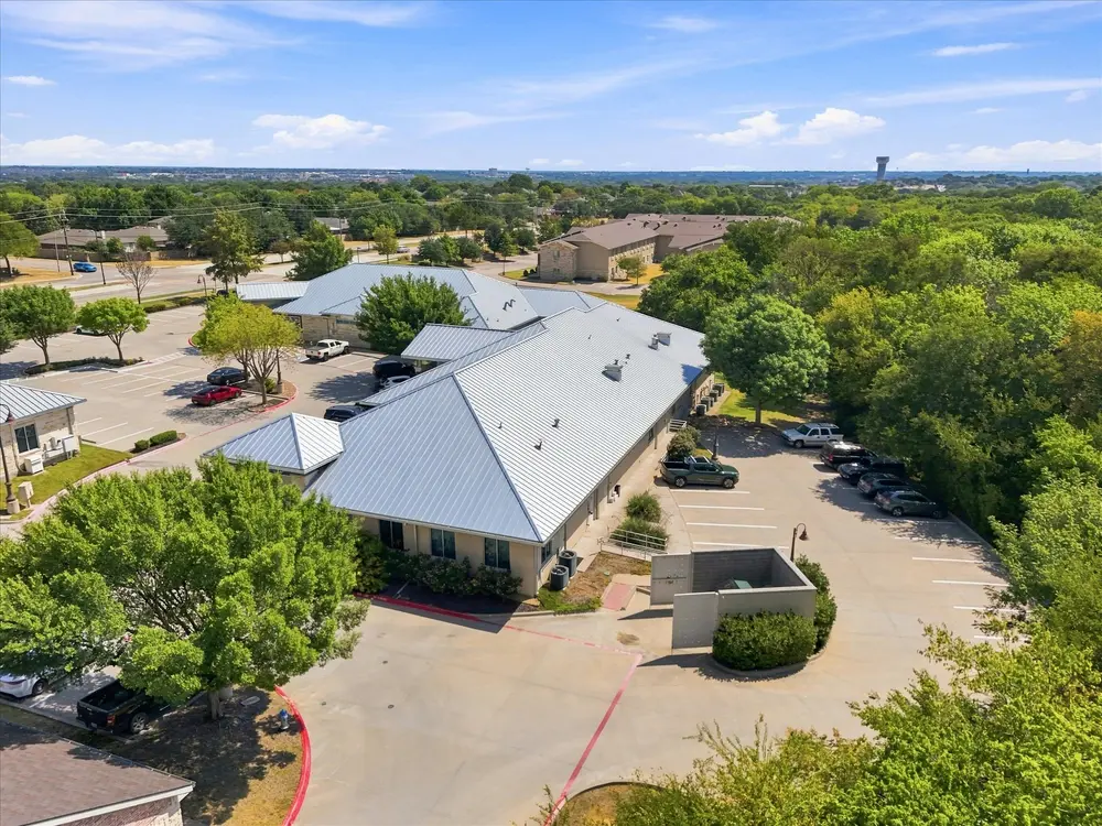 Angled aerial of a metal-roof office building, its parking lot and the tree line behind it
